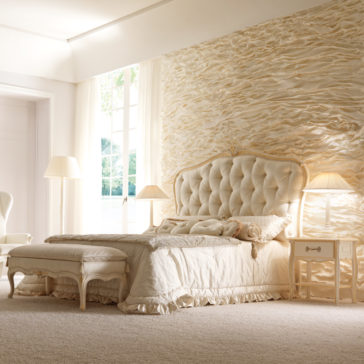 Italian Ivory And Gold Luxury Reproduction Bed A luxurious bedroom showcases an Italian Ivory And Gold Luxury Reproduction Bed with an elegant, tufted headboard, accompanied by a matching bench at its foot. The space is adorned in soft, neutral tones featuring textured, wavy wall accents and large windows that flood the room with natural light. Completing the serene setting are a nightstand with a stylish lamp and a cozy chair.