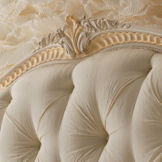 Italian Ivory And Gold Luxury Reproduction Bed Close-up of an ornate, cream-colored headboard with intricate carvings and tufted fabric. The detailed carvings feature elegant floral patterns, reminiscent of the Italian Ivory And Gold Luxury Reproduction Bed, adding a touch of sophistication to the headboard's design.