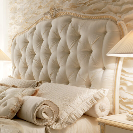 Italian Ivory And Gold Luxury Reproduction Bed A luxurious bedroom showcases the Italian Ivory and Gold Luxury Reproduction Bed, complete with an ornate, tufted headboard in cream and elegant detailing. Plush matching pillows and a rolled cushion adorn the bed. The headboard is set against a textured, wave-like wall, flanked by two beige lamps.