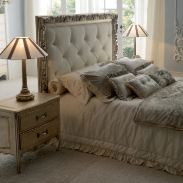 A cozy bedroom features an Italian Reproduction Button Upholstered Bed adorned with beige and gray pillows and bedding. Two bedside tables with decorative lamps flank the bed. Natural light streams through large windows with partially drawn white curtains, illuminating the serene space.