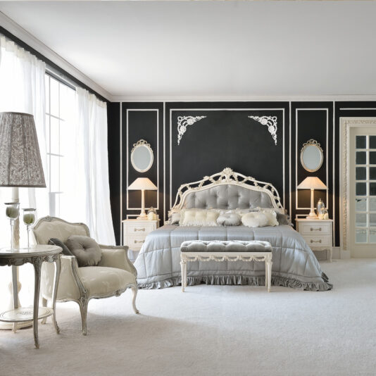An elegant bedroom features the Classic Style Italian Button Upholstered Bed, showcasing a tufted gray headboard and ruffled bedding. The room has tall windows with sheer curtains, a plush armchair, ornate side tables with lamps, wall mirrors, and a black accent wall with intricate white molding.