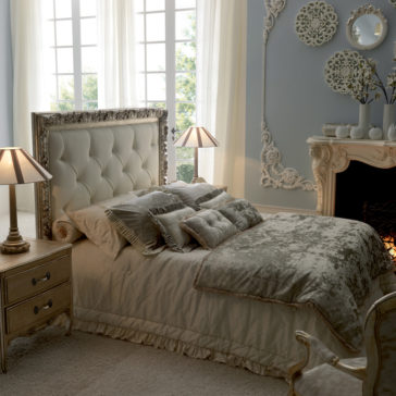 A luxurious, elegant bedroom showcasing the Italian Reproduction Button Upholstered Bed, adorned with plush velvet-printed bedding and multiple accent pillows. Ornate nightstands with table lamps flank the bed, while a fireplace adorned with decorative mirrors and vases graces the mantle.