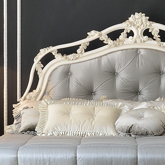 A close-up view of the Classic Style Italian Button Upholstered Bed showcases its luxurious, tufted silver headboard adorned with intricate floral patterns. The bedding ensemble features silver and cream pillows paired with a matching comforter, creating an elegant and sophisticated aesthetic.