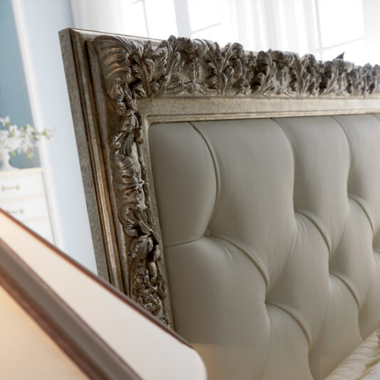 Italian Reproduction Button Upholstered Bed Close-up of the Italian Reproduction Button Upholstered Bed in a bedroom. The headboard showcases cream-colored, tufted leather and an intricately carved, antique frame with a metallic finish. Soft, natural light filters through curtains in the background.