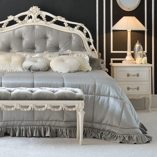 A beautifully decorated bedroom featuring the Classic Style Italian Button Upholstered Bed with a large, tufted headboard adorned with intricate carvings. The bed is draped in a matching grey satin bedspread accompanied by multiple decorative pillows. An elegant bench sits at the foot of the bed, while a classic bedside table with a lamp completes the look. Chic dark accent walls perfectly complement this luxurious setup.