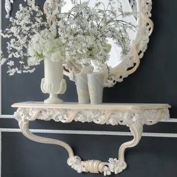 An Italian Rose and Ribbon Reproduction Wall Fixing Console Table features a decorative wall shelf with intricate carvings and an ornate mirror. On the shelf, glass vases filled with white flowers and small branches complete the lovely arrangement. This setup rests against a dark wall with white trim, creating an elegant and classic look.