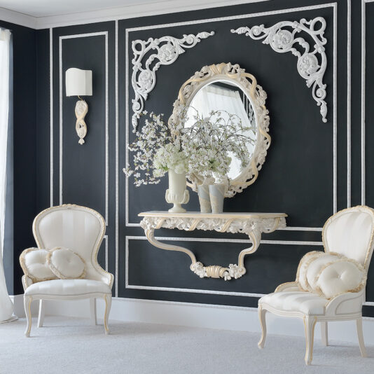 An elegant room showcases a striking black wall, highlighted by intricate white decorative moldings. A baroque-style mirror complements the space, while an Italian Rose and Ribbon Reproduction Wall Fixing Console Table elegantly displays floral arrangements. Two upholstered armchairs with heart-shaped pillows add cozy seating, and light streams in from a tall window on the left.
