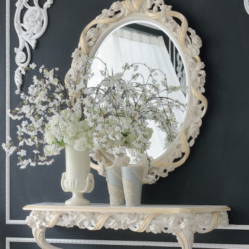 An elegant room featuring a large, ornate round mirror with a decorative frame hung on a dark wall. Below it stands the Italian Rose and Ribbon Reproduction Wall Fixing Console Table, adorned with a white vase of blooming white flowers and two smaller gray vases, creating a sophisticated and classic look.