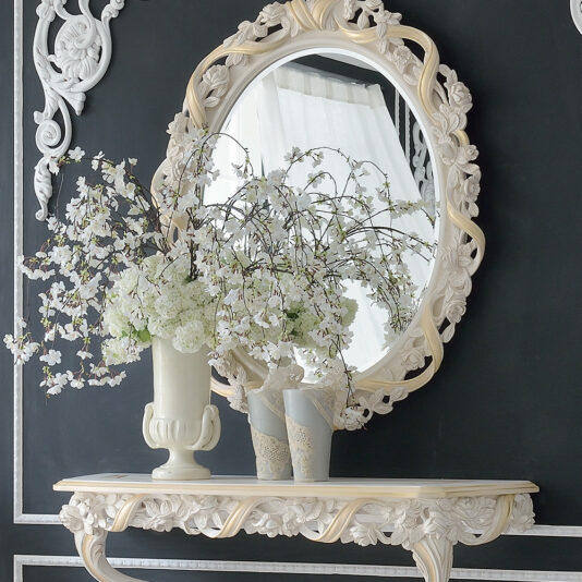 An elegant room featuring a large, ornate round mirror with a decorative frame hung on a dark wall. Below it stands the Italian Rose and Ribbon Reproduction Wall Fixing Console Table, adorned with a white vase of blooming white flowers and two smaller gray vases, creating a sophisticated and classic look.