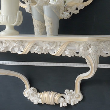 A close-up of the Italian Rose and Ribbon Reproduction Wall Fixing Console Table, featuring an ornate cream-colored design with intricate floral carvings, supporting a framed mirror above. The console displays a small vase with white flowers and a decorative urn, set against a dark wall.