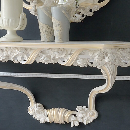 A close-up of the Italian Rose and Ribbon Reproduction Wall Fixing Console Table, featuring an ornate cream-colored design with intricate floral carvings, supporting a framed mirror above. The console displays a small vase with white flowers and a decorative urn, set against a dark wall.
