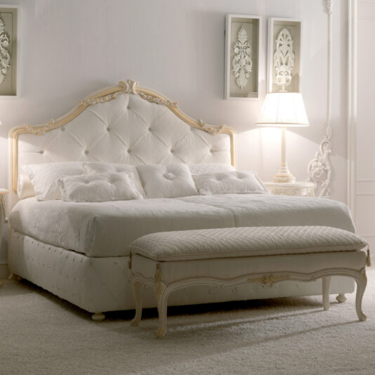 A luxurious bedroom featuring the Italian Rose Detail Luxury Bed, a plush, tufted white bed with an ornate rose detail headboard and a matching bench at the foot. Textured white pillows and a quilted comforter adorn the bed. Elegant white decor, including a lamp and framed artwork, complete the serene, sophisticated look.