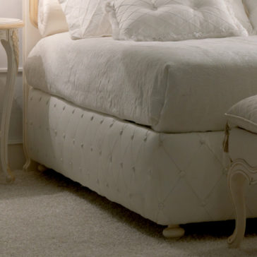 Italian Rose Detail Luxury Bed An Italian Rose Detail Luxury Bed, featuring a plush, white frame with an exquisitely quilted headboard adorned with Italian rose details and matching pillows, sits in the corner of the room. Next to it is a white bedside table with gracefully curved legs and a chic lamp. The room exudes a cozy ambiance with its soft, neutral color palette.
