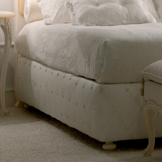 An Italian Rose Detail Luxury Bed, featuring a plush, white frame with an exquisitely quilted headboard adorned with Italian rose details and matching pillows, sits in the corner of the room. Next to it is a white bedside table with gracefully curved legs and a chic lamp. The room exudes a cozy ambiance with its soft, neutral color palette.