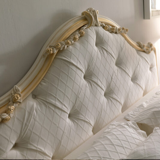 Here is a close-up view of the Italian Rose Detail Luxury Bed, featuring an elegant cream-colored tufted headboard adorned with intricate Italian rose details and a quilted fabric design. The luxury bed is partially covered with a white, textured bedspread as sunlight streams in from a nearby window.