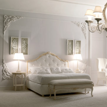 Italian Rose Detail Luxury Bed A luxurious bedroom with an ornate design featuring the Italian Rose Detail Luxury Bed adorned with white tufted upholstery, complemented by a matching bench and two nightstands with lamps. Elegant wall moldings grace the walls along with framed decorative art pieces, while a chandelier with multiple lamps illuminates the space from above.