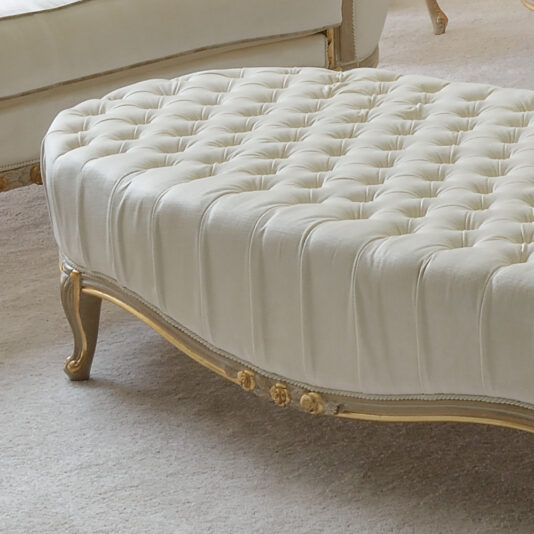 A close-up image of a tufted, cream-colored Louis Reproduction Button Upholstered Coffee Table with carved wooden legs and gold trim. This elegant piece boasts a sophisticated style, complemented by a matching sofa visible in the background. Both items rest on a light-colored carpeted floor, exuding timeless sophistication.