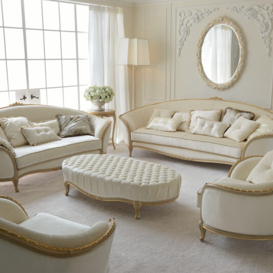 A luxurious and elegant living room with ornate white and gold furniture, including two sofas, two armchairs, and a tufted ottoman. A large mirror with a decorative frame hangs on the wall, while a Louis Reproduction Button Upholstered Coffee Table sits beside a tall lamp near a vase of white flowers by the window.