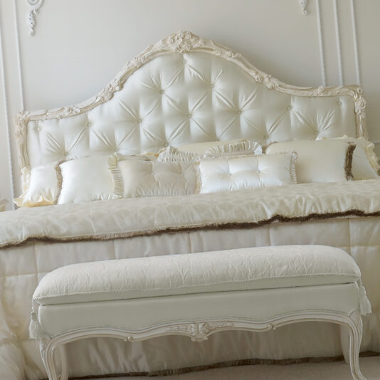 A Luxurious Classic Italian Designer Button Upholstered Bed features an elegant, tufted headboard, adorned with decorative pillows and an ornate quilt. A matching button-upholstered bench with intricate carvings sits at the foot of this exquisite bed. The predominantly white decor with subtle textures creates a serene ambiance.
