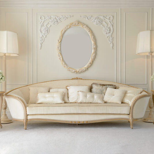 A luxurious living room features the exquisite Luxury Italian Ivory Louis Reproduction Sofa, adorned with multiple plush pillows. Above the sofa hangs an ornate, oval mirror with intricate carvings. The room is enriched with elegant wall moldings and flanked on either side by tall, decorative floor lamps.