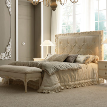 A luxurious bedroom featuring a large Luxury Ornate Button Upholstered Bed adorned with beige bedding and multiple pillows. An upholstered bench sits at the foot of the bed. Two nightstands with table lamps flank the ornate bed, and a large window with sheer curtains lets in natural light.