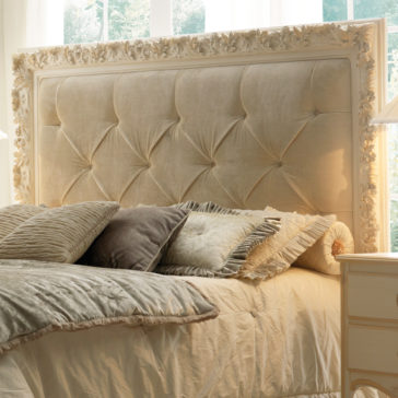 An Opulent Italian Swirl Gold Button Upholstered Bed with a tufted headboard and several pillows in varying shades of beige, brown, and gray. The bed is covered with a cream-colored blanket, and the scene is warmly lit by daylight streaming through large windows in the background.