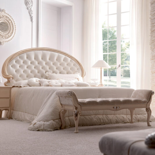 An elegant bedroom showcases the Opulent Italian Swirl Gold Button Upholstered Bed, adorned with plush white bedding and complemented by a cushioned bench at the foot. Large windows with sheer curtains invite natural light to bathe the serene space. A bedside table featuring a lamp and an ornate mirror adds charm to the room.