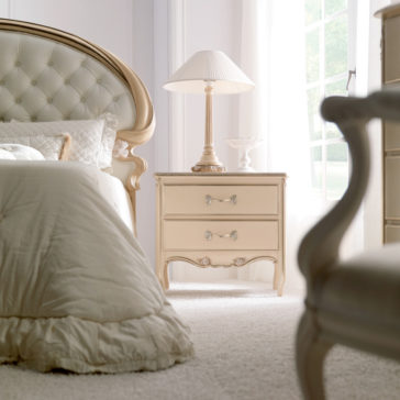 Opulent Italian Swirl Gold Button Upholstered Bed A luxurious bedroom featuring an Opulent Italian Swirl Gold Button Upholstered Bed with beige bedding, a wooden bedside table with two drawers, and a classic table lamp. An armchair is partially visible in the foreground, and the room is brightly lit by natural light from a nearby window.