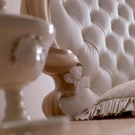 Opulent Italian Swirl Gold Button Upholstered Bed Close-up of the Opulent Italian Swirl Gold Button Upholstered Bed, showcasing its luxurious, tufted white headboard with elegant wooden detailing. An ornate translucent floral accent is attached to the wood frame. Part of an opulent Italian swirl ceramic vase is blurred in the foreground, and fringed bedding on the bed is visible.