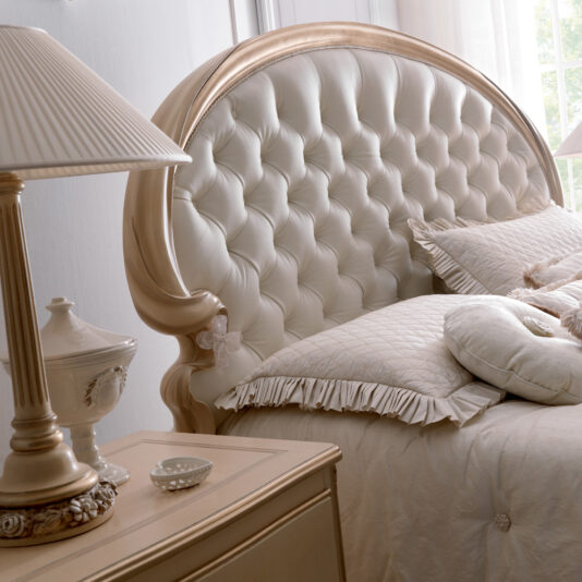 Opulent Italian Swirl Gold Button Upholstered Bed A luxurious bedroom showcases the Opulent Italian Swirl Gold Button Upholstered Bed, adorned with quilted bedding and decorative pillows. Adjacent to the bed is a stylish nightstand holding a classic lamp, a small ceramic dish, and a decorative item, all in a soft, elegant color scheme.