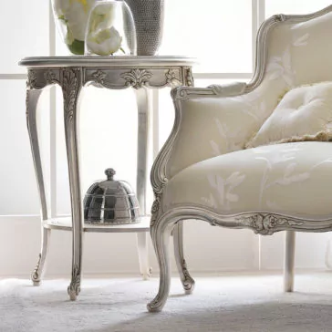 Ornate Silver Italian Side Table An elegant living room scene featuring a vintage, floral-patterned armchair with intricate wooden carvings. Beside it, the Ornate Silver Italian Side Table holds a glass vase with white flowers and a silver serving dish on the lower shelf. Sunlight filters in softly from the background.
