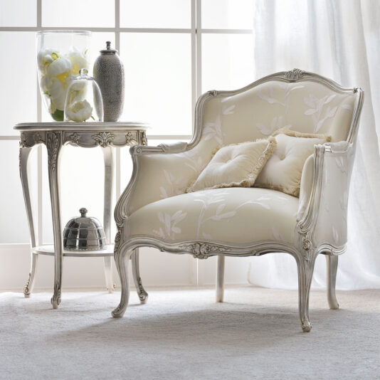 A timeless armchair adorned with floral upholstery, paired with two plush pillows, is placed next to the Ornate Silver Italian Side Table. The table holds a ceramic vase filled with white flowers, a decorative jar, and a small lidded dish. This charming arrangement sits beside a large, light-filled window.