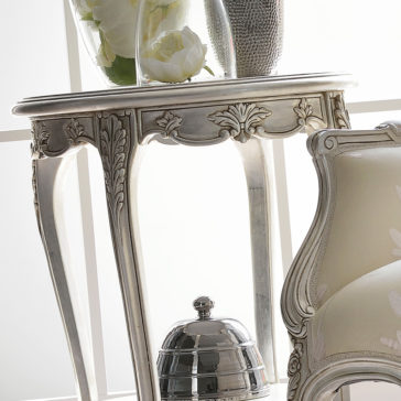 Ornate Silver Italian Side Table A close-up of an elegant corner featuring the Ornate Silver Italian Side Table, which boasts intricately carved, curved legs. On the table, there's a vase with white flowers and a silver decorative object. Beside the table is a plush, light-colored upholstered chair.