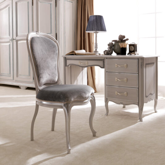 An elegant room featuring a vintage-style desk with curved legs, three drawers, and decorative handles. Resting on the desk are a lamp, clock, and some small decorative items. Positioned in front of the desk is a sophisticated gray upholstered Reproduction Designer Italian Chair with silver trim.