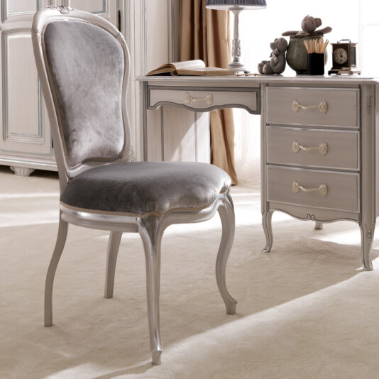 In a vintage-styled room, the Reproduction Designer Italian Chair, upholstered in plush gray velvet and exuding timeless elegance, stands out with its high, curved back and ornate wooden legs. Next to it is a matching wooden desk showcasing intricate carved details, gold handles, and drawers adorned with various decorative items.