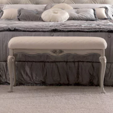 Reproduction Italian Bedroom Bench An elegant bedroom boasts a spacious, plush bed adorned with a tufted headboard and luxurious silver-grey bedding. Positioned in front of the bed is an ornate cream-colored Reproduction Italian Bedroom Bench, featuring intricately carved wooden legs and a cushioned seat. The room is gently carpeted for added softness.