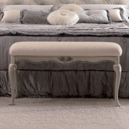 An elegant bedroom boasts a spacious, plush bed adorned with a tufted headboard and luxurious silver-grey bedding. Positioned in front of the bed is an ornate cream-colored Reproduction Italian Bedroom Bench, featuring intricately carved wooden legs and a cushioned seat. The room is gently carpeted for added softness.