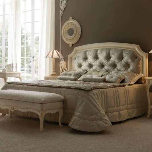 Elegant bedroom featuring the Reproduction Italian Button Upholstered Bed with a plush, tufted headboard in a champagne hue. The bed is adorned with matching bedding and pillows. Ornate side tables with classic lamps flank the bed, while a decorative round mirror hangs above the headboard. A bench is placed at the foot of the bed.
