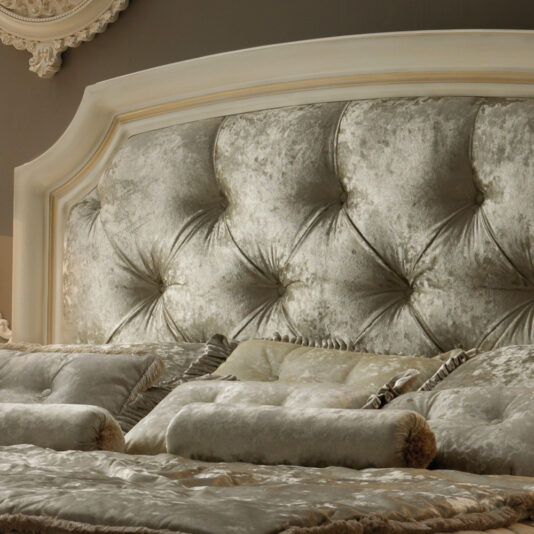 The Reproduction Italian Button Upholstered Bed is a luxurious and plush bed adorned with a tufted, silver headboard and matching silver bedding. It includes several cushioned pillows and bolsters in coordinated silver fabric, creating an elegant and opulent appearance.