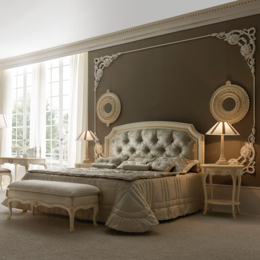 A luxurious bedroom features a Reproduction Italian Button Upholstered Bed with a tufted headboard, cream-colored bedding, and two bedside tables with matching lamps. The room boasts ornate wall decor and large floor-to-ceiling windows that let in ample natural light.