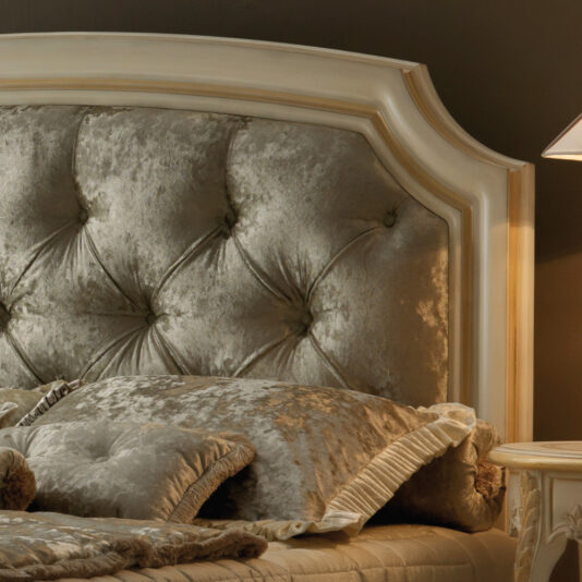 The Reproduction Italian Button Upholstered Bed, featuring luxurious beige and cream-colored upholstery, showcases a tufted headboard with elegant curves and detailing. Plush, silky pillows and bedding enhance the comfort, while a softly lit lamp on the right casts a warm glow over the cozy setting.