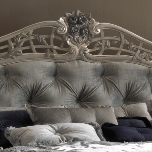 The Trailing Roses Italian Button Upholstered Designer Bed features a luxurious, tufted headboard with intricate floral carvings at the top. Adorned with an array of plush pillows in various shades of grey and navy blue, this sophisticated bed exudes elegance.