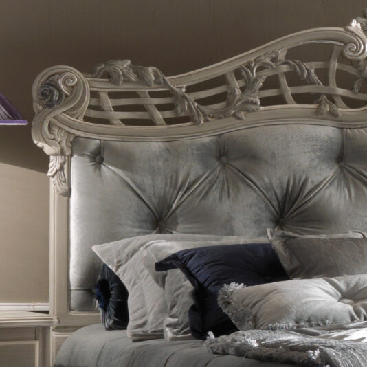 The Trailing Roses Italian Button Upholstered Designer Bed features a luxurious and ornate gray tufted headboard with intricate wood carvings. At the head of the neatly made bed, it is adorned with soft, elegant pillows in shades of white, gray, and blue, and a quilted comforter, enhancing the room's sophisticated ambiance.