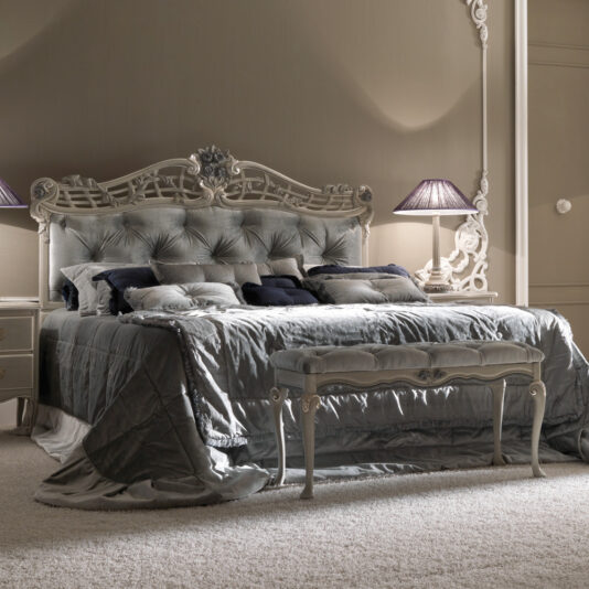 A luxurious bedroom showcases the Trailing Roses Italian Button Upholstered Designer Bed, featuring an ornate headboard and footstool. The bed is draped in plush gray bedding with numerous pillows, and elegant nightstands with matching lamps flank it. The walls are adorned with intricate moldings, enhancing the sophisticated ambiance.