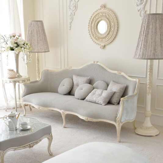 A luxurious living room showcases the Venetian Style Soft Grey Designer Sofa, adorned with several decorative pillows. Ornate floor lamps flank the sofa, and a round, ornate wall mirror is positioned above it, infusing the space with Venetian charm. A side table featuring a floral arrangement adds a fresh touch.