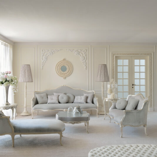 Elegant living room with light gray and white decor. Features classic furniture, including a Venetian Style Soft Grey Designer Sofa, armchairs, and an ottoman. Intricate moldings and a round mirror adorn the walls. Two tall lamps stand on either side of the sofa, and large windows bring in natural light.