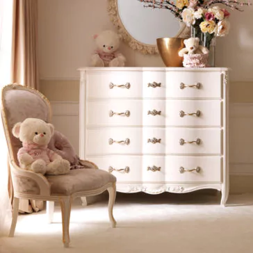 A plush chair adorned with a teddy bear and a pink pillow is placed next to the Designer Italian Chest of Drawers, featuring ornate floral handles. On top of the dresser sits a teddy bear and a vase filled with flowers, while a round decorative mirror hangs on the wall above it. Sunlight streams in from the left.