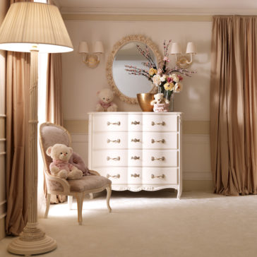 A cozy, elegant bedroom with a vintage feel features the Designer Italian Chest of Drawers adorned with flowers and a round mirror above it. A plush teddy bear sits on an ornate chair next to a floor lamp. The room is decorated in soft beige and white tones with flowing curtains.