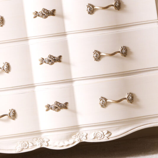 A close-up of the Designer Italian Chest of Drawers showcases three drawers adorned with intricate gold handles. The chest boasts elegant carvings and floral details, enhancing its vintage charm.