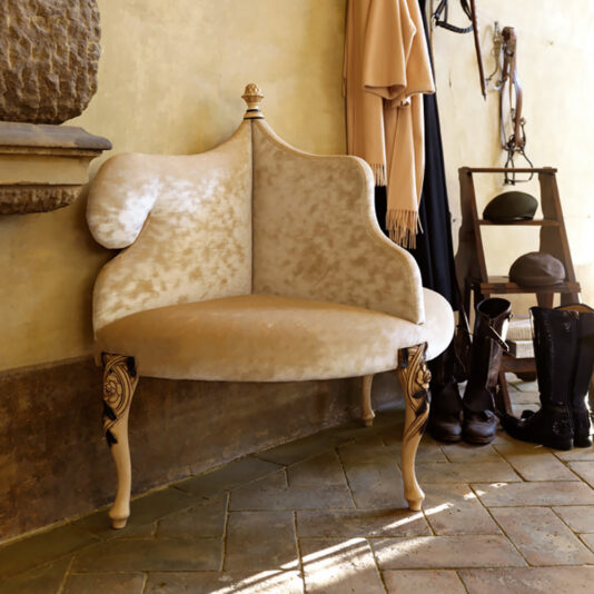 A Designer Italian Entry Bench, featuring ornate cream-colored carvings and seating for two, stands in a room with tan walls and a tiled floor. Surrounding it are equestrian items such as boots, helmets, and riding gear on wooden shelves. A hanging coat is partially visible.